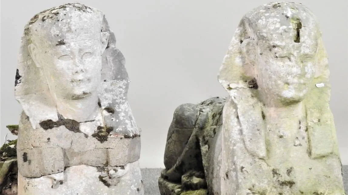 Dos estatuas de esfinges egipcias de piedra desgastadas y cubiertas de liquen, enfrentadas entre sí.