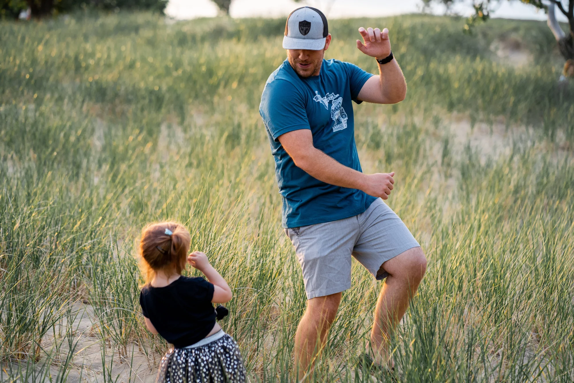 Un hombre con gorra y camiseta azul baila y juega con una niña pequeña entre hierbas altas en dunas de arena.