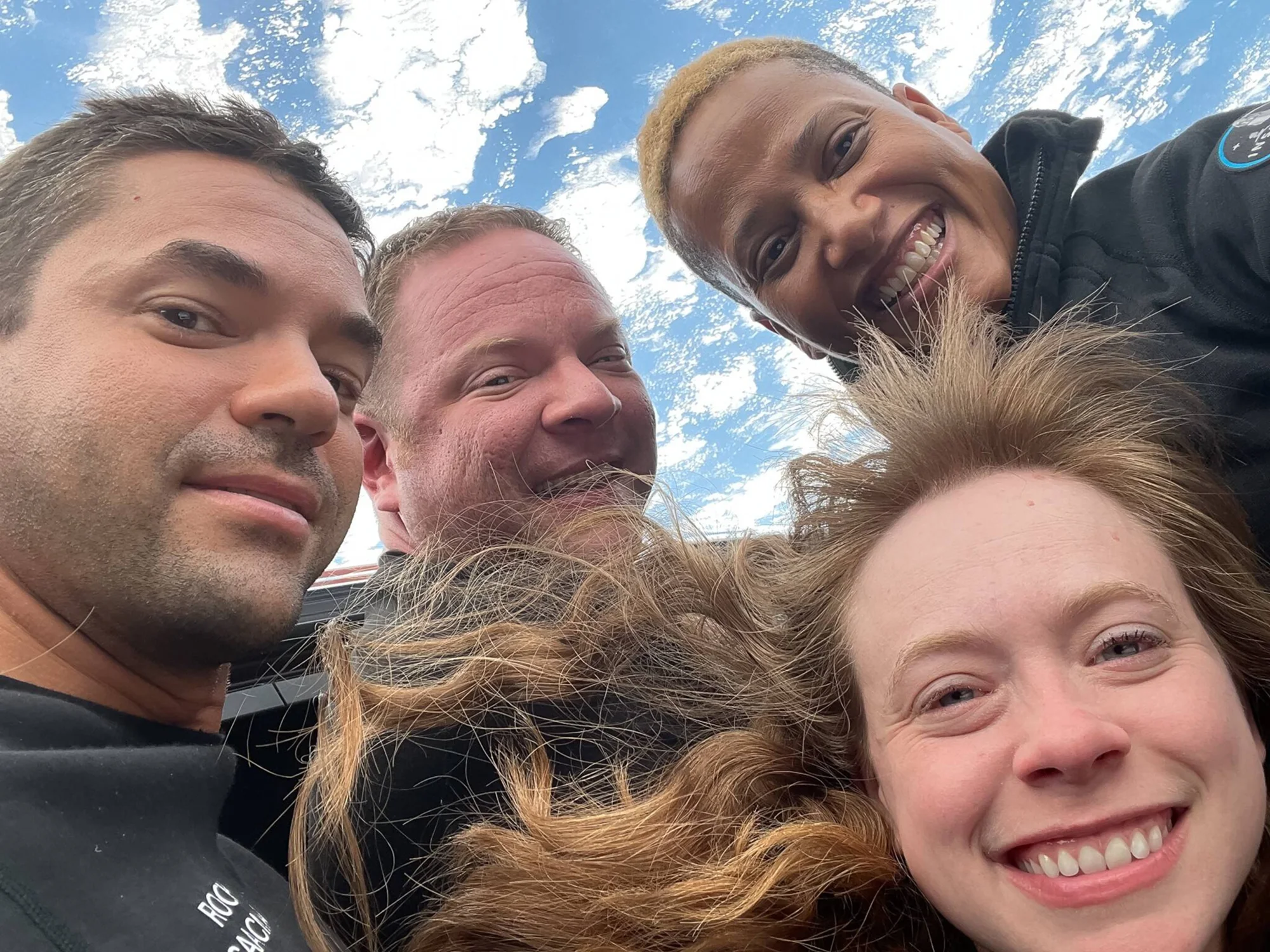 Cuatro personas sonríen juntas en una selfie tomada al aire libre bajo un cielo azul con nubes.