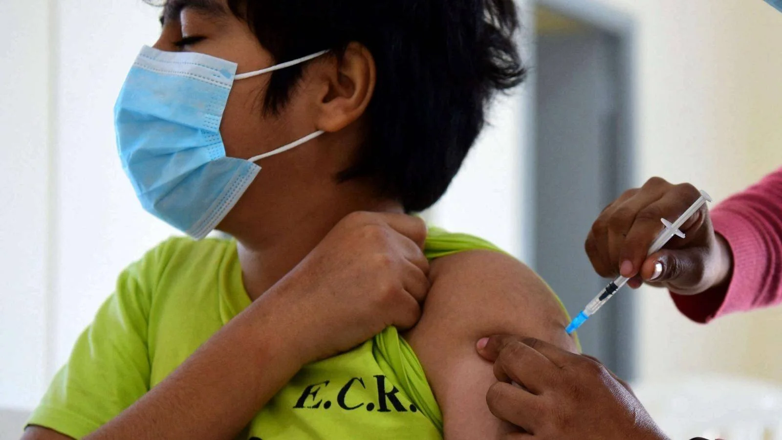 Un niño con mascarilla quirúrgica recibe una inyección en el brazo por parte de un profesional de salud.