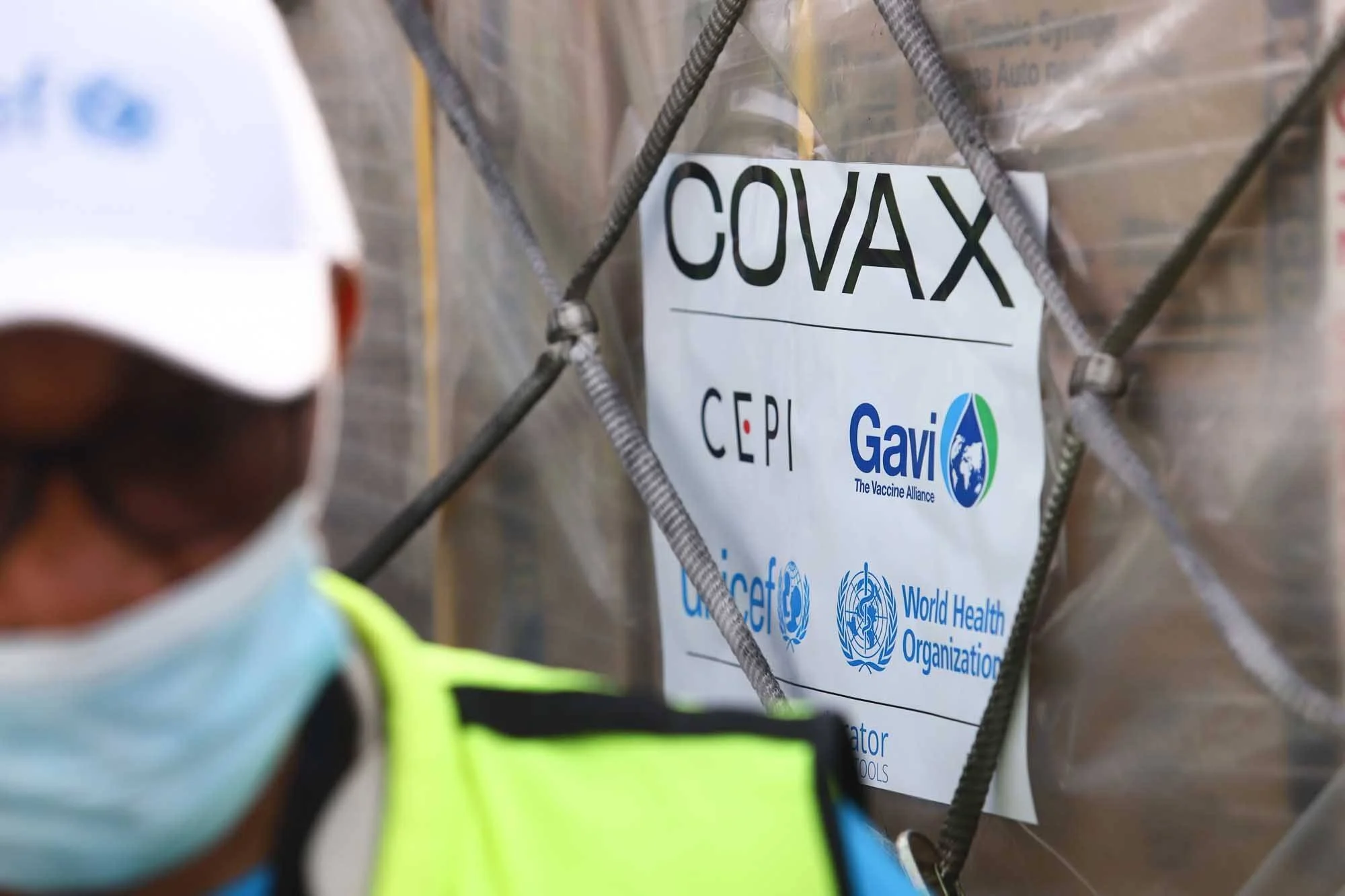 Trabajador con mascarilla junto a un pallet identificado con el logo COVAX, CEPI, Gavi, UNICEF y la OMS.