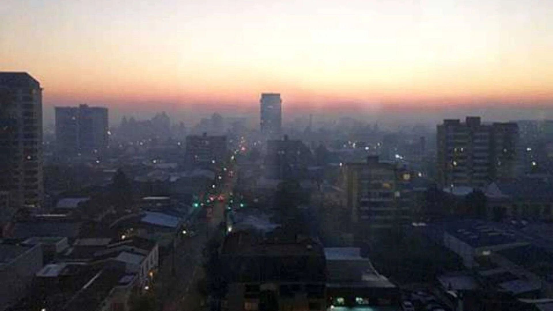 Panorámica urbana al amanecer con una densa capa de smog que cubre los edificios y una avenida iluminada al centro.