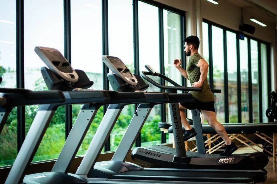 Un hombre con remera verde corre en una cinta Assault Fitness AirRunner en un gimnasio iluminado con ventanales.