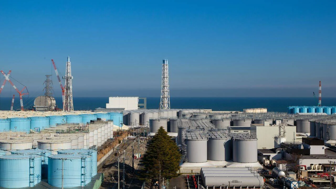Cientos de tanques de almacenamiento de agua azules y grises en una planta nuclear ubicada frente al océano, con grúas y torres visibles al fondo.
