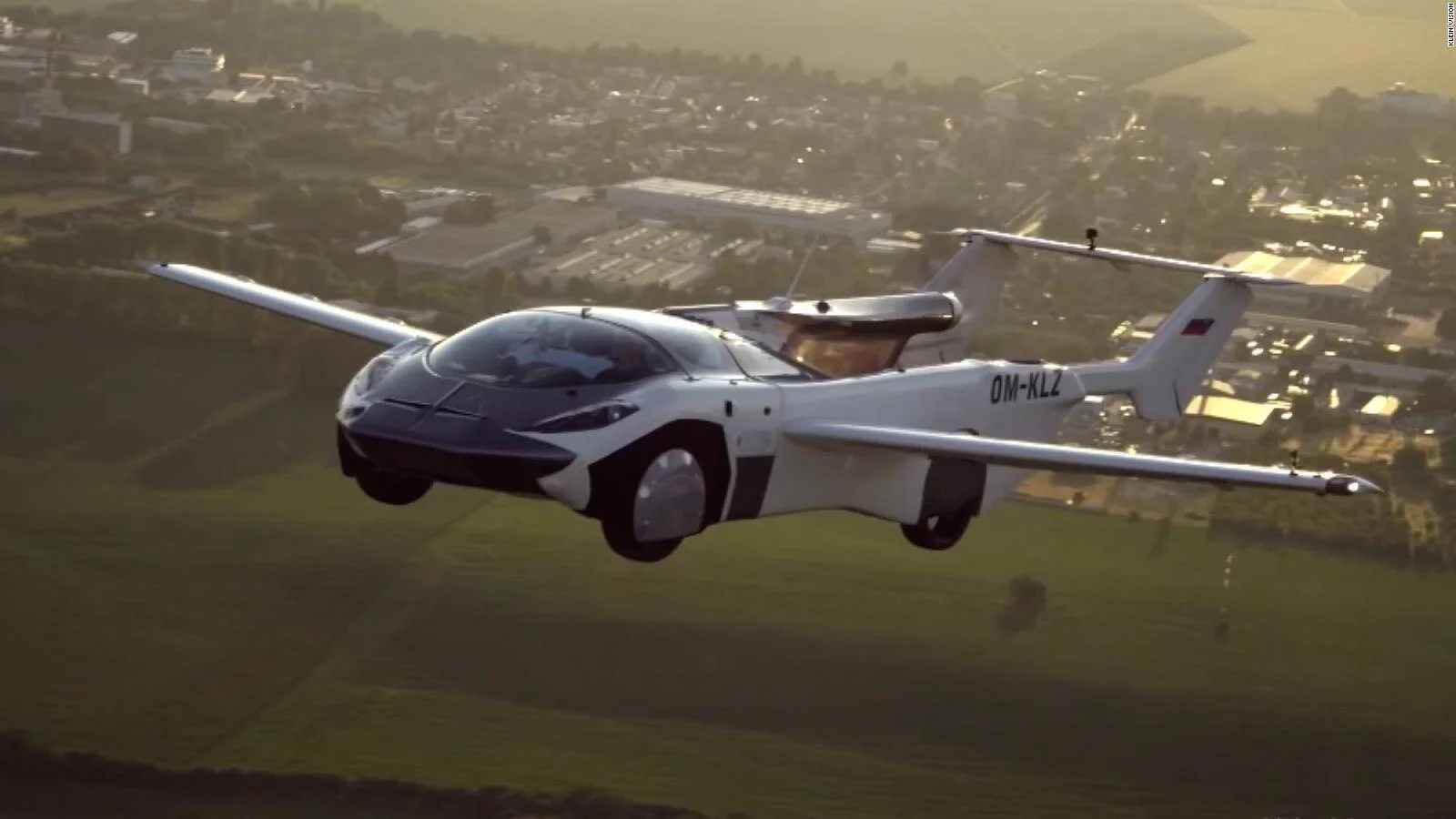 Vehículo híbrido auto-avión con matrícula OM-KLZ volando sobre paisaje urbano y rural.