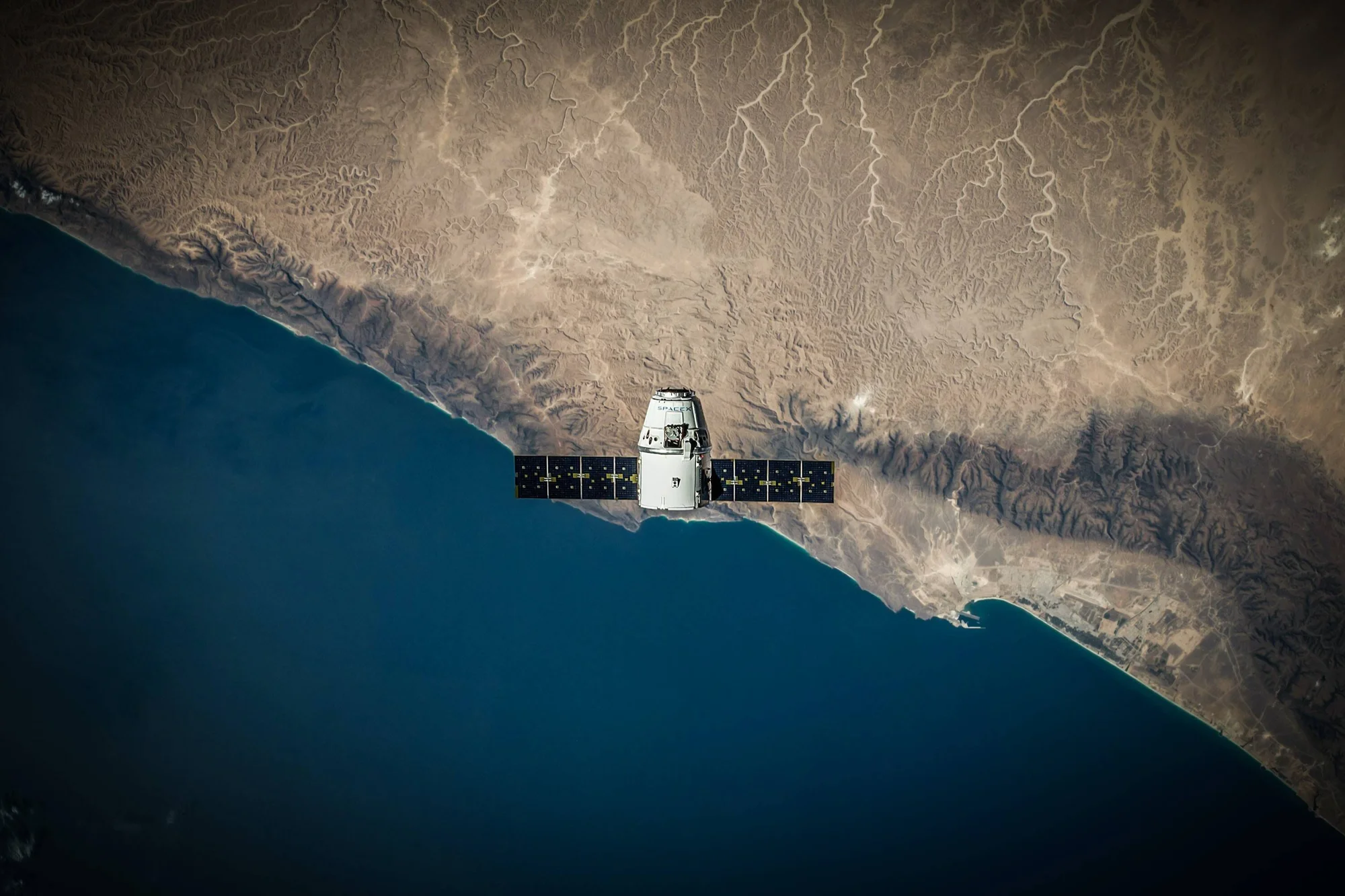 Cápsula espacial SpaceX Dragon con paneles solares desplegados sobrevolando una costa desértica vista desde el espacio.