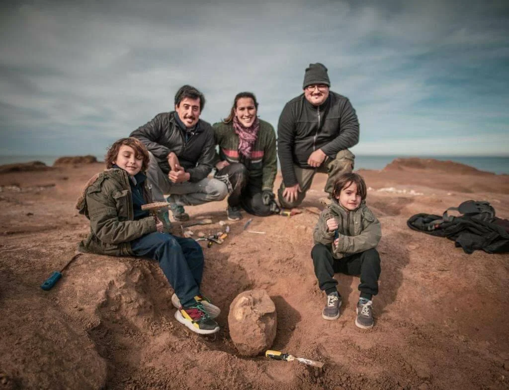 Tres adultos y dos niños posan sonrientes junto a un fósil recién excavado en un yacimiento paleontológico a cielo abierto.
