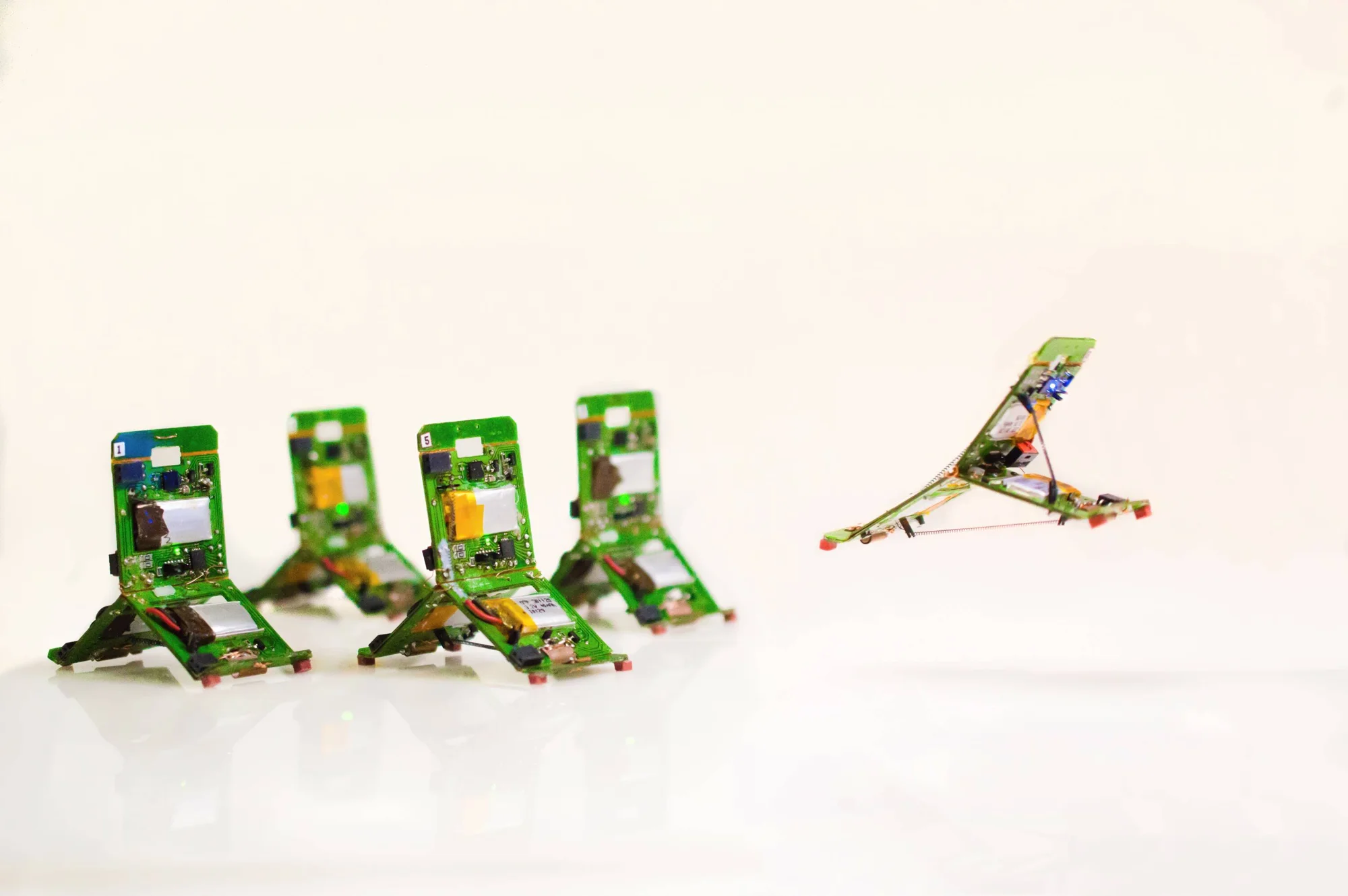 Cuatro pequeños robots con circuitos impresos verdes sobre el suelo y uno de ellos volando con alas desplegadas.