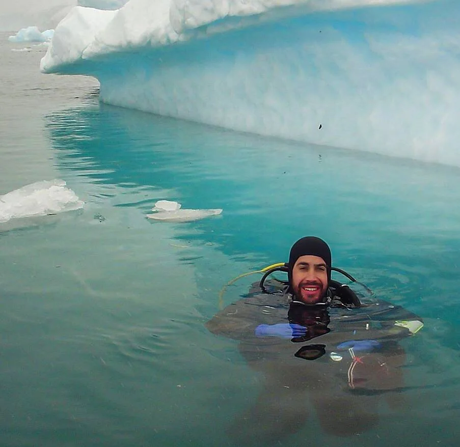 Un buzo con traje de neopreno sonríe en aguas heladas rodeado de hielo y un gran iceberg azul al fondo.