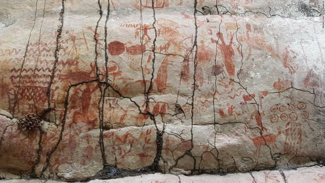 Pared de roca con numerosas pinturas rupestres en ocre rojizo representando figuras humanas, animales y símbolos geométricos.