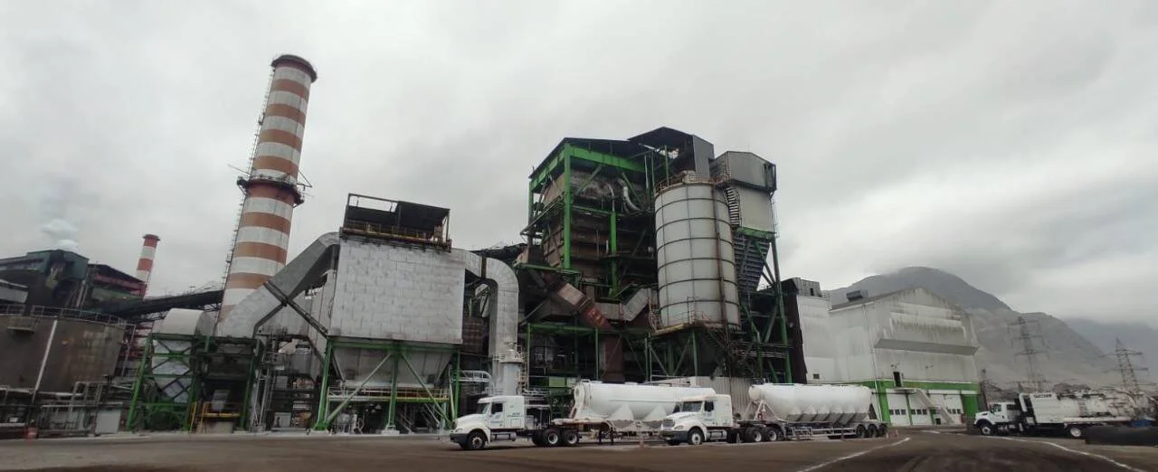 Complejo industrial de gran escala con chimeneas a rayas, estructuras metálicas verdes y camiones cisterna estacionados al frente.