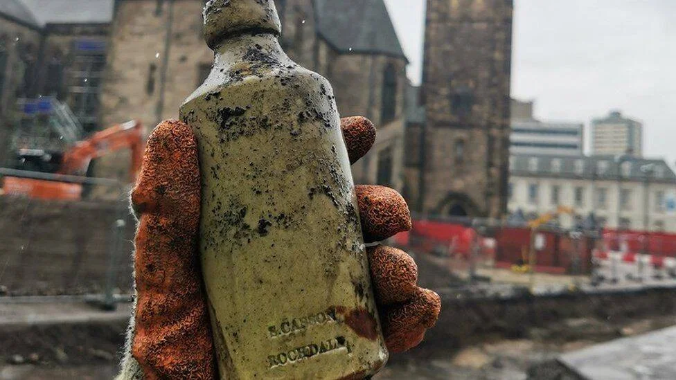 Mano con guante sostiene una botella antigua con inscripción 'B. Casson Rochdale', encontrada en una excavación urbana junto a una iglesia de piedra.