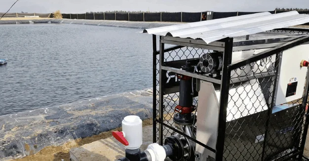 Sistema de bombas y tuberías instalado al borde de una laguna de tratamiento de agua, con estructura metálica y techo protector.