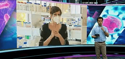 Presentador de televisión junto a una pantalla grande que muestra a una mujer con mascarilla en un laboratorio, con gráficos de virus al fondo.