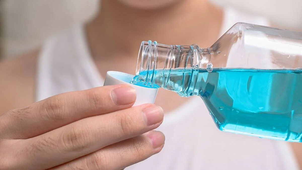 Manos vertiendo enjuague bucal líquido azul desde una botella transparente hacia un pequeño vaso dosificador blanco.