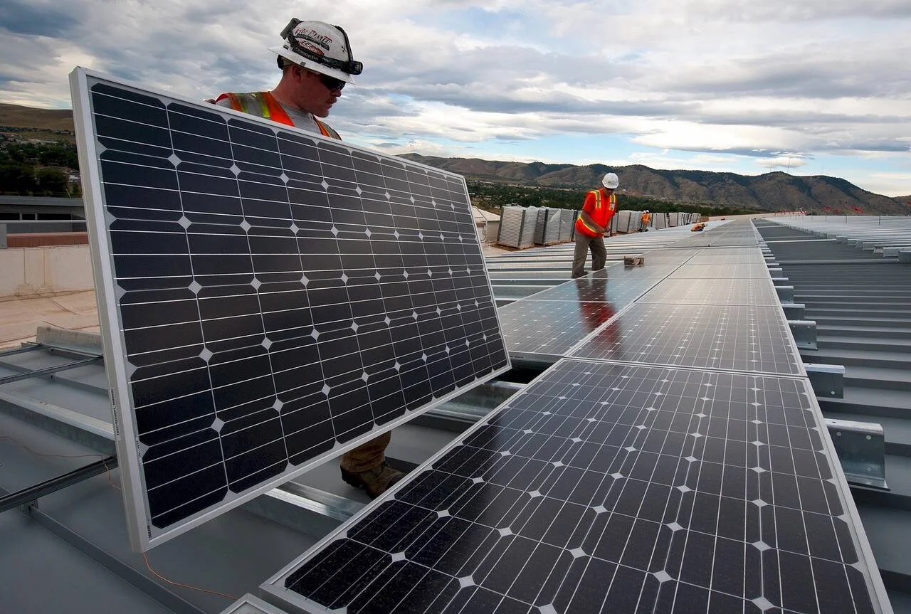 Dos operarios con cascos y chalecos reflectantes instalan paneles solares fotovoltaicos sobre una estructura metálica en un techo, con montañas al fondo.