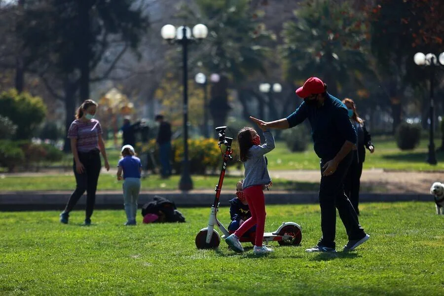 Un adulto con mascarilla ayuda a una niña junto a un scooter eléctrico en un parque, mientras otras personas pasean al fondo.
