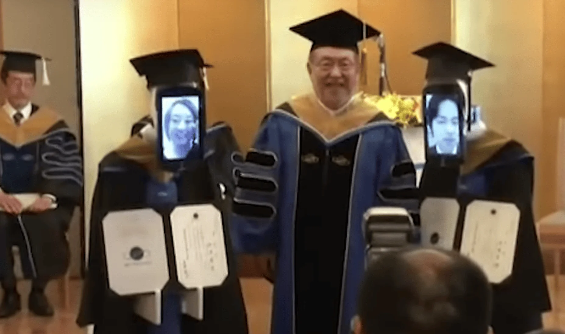 Robots con pantallas que muestran rostros de estudiantes remotos, vestidos con toga y birrete, reciben diplomas en una ceremonia de graduación.