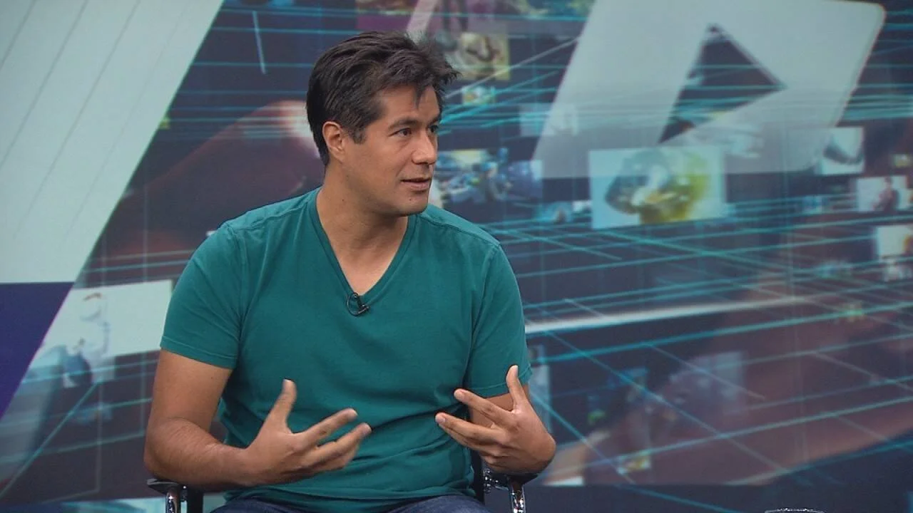 Hombre con remera verde gesticula mientras habla sentado en un estudio de televisión con fondo tecnológico.
