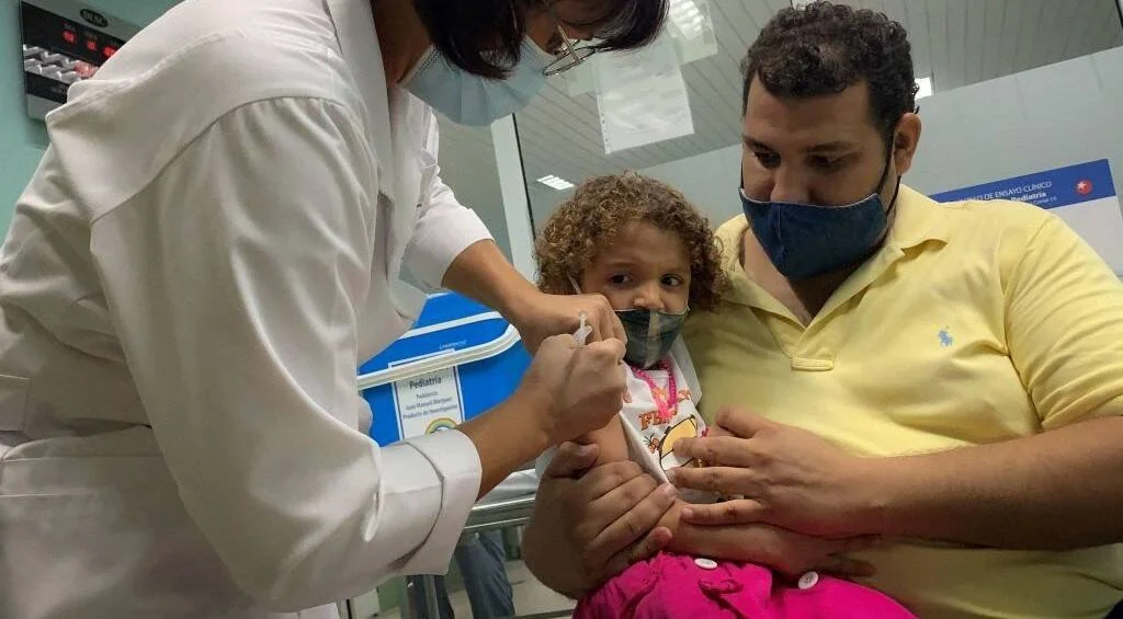 Una enfermera aplica una vacuna en el brazo de una niña pequeña sostenida por un adulto con mascarilla en un centro de salud.
