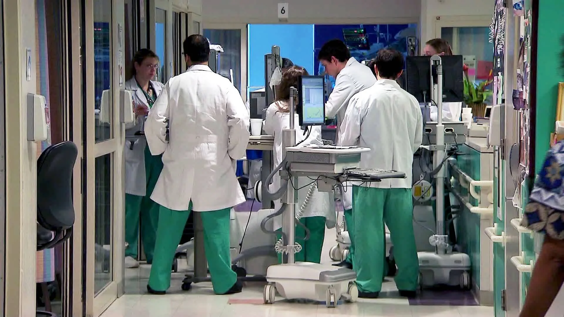 Varios médicos con batas blancas y pijamas quirúrgicos verdes consultan computadoras en el pasillo de un hospital.