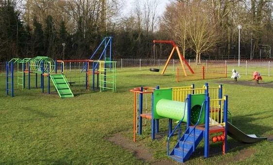 Parque infantil con estructuras de colores, tobogán, columpio y túnel sobre césped verde rodeado de árboles.
