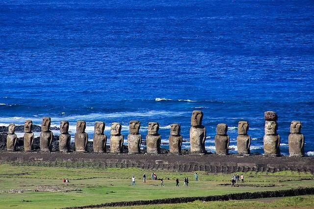 Fila de quince estatuas moái frente al océano Pacífico en Isla de Pascua, con turistas al pie de la plataforma.