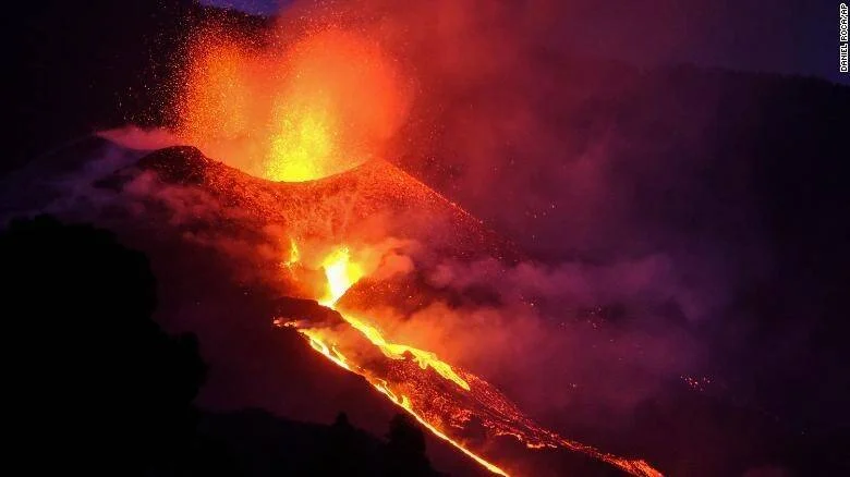 Volcán en erupción expulsando lava incandescente y una gran columna de fuego durante la noche, con ríos de lava descendiendo por su ladera.