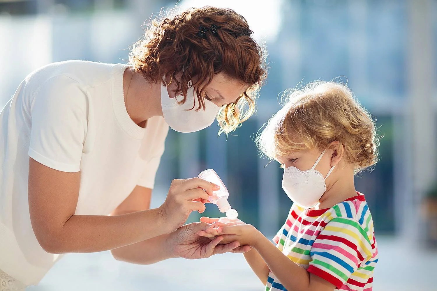 Una mujer con mascarilla aplica gel desinfectante en las manos de un niño pequeño también enmascarado, al aire libre.