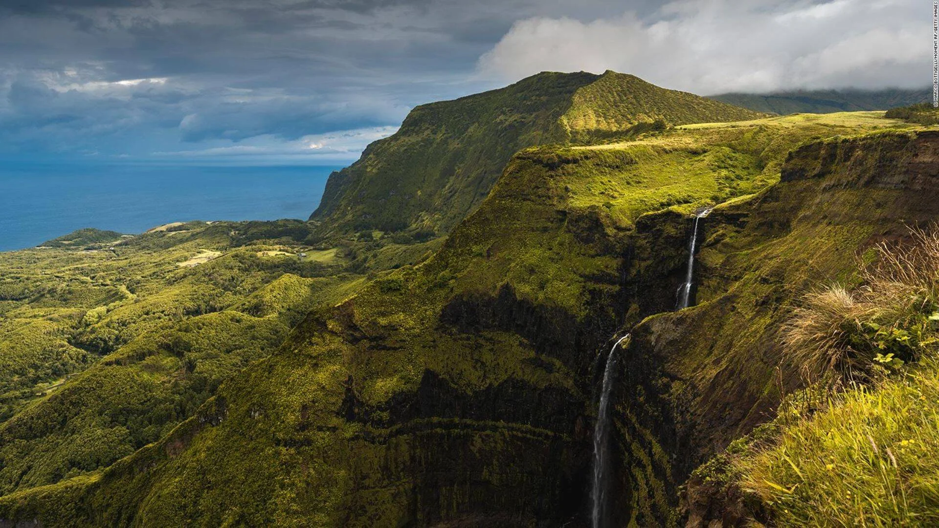 Cascada cae por un acantilado cubierto de vegetación exuberante, con un valle verde y el océano azul al fondo bajo cielo nublado.