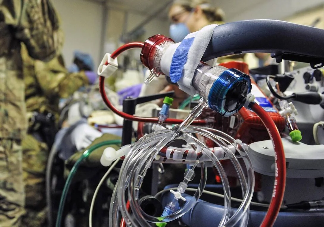 Primer plano de un equipo de oxigenación por membrana extracorpórea (ECMO) con tubos y circuitos, con personal médico y militar al fondo.