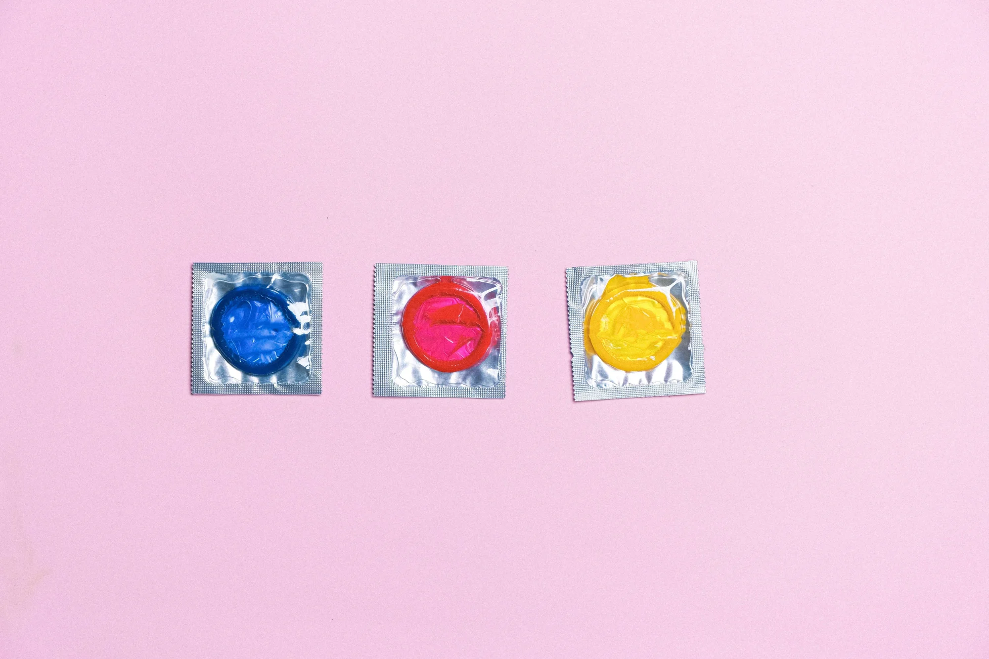 Tres preservativos en sus envases plateados, de colores azul, rojo y amarillo, alineados sobre fondo rosa.