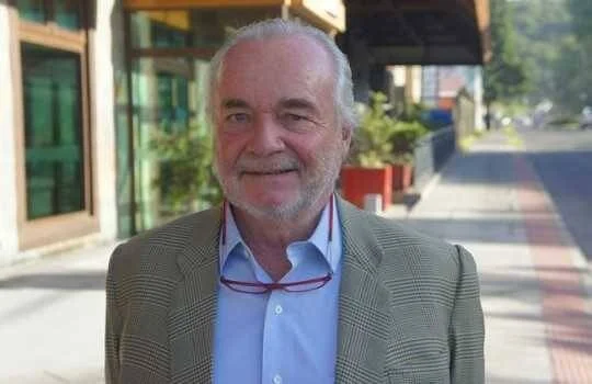 Hombre mayor de cabello y barba blancos, con blazer gris y camisa azul, posando en una calle soleada.