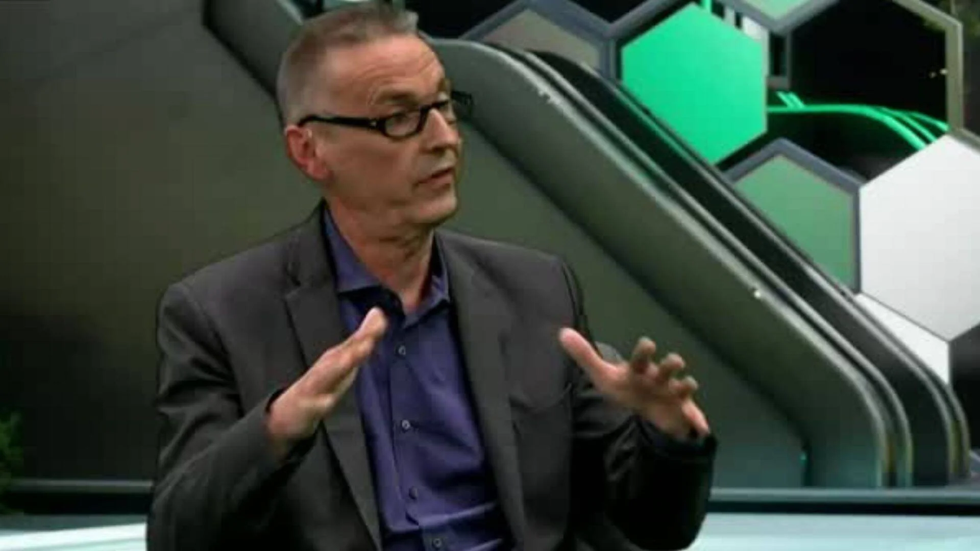 Hombre con gafas y saco oscuro gesticula con ambas manos mientras habla en un estudio de televisión con decorado hexagonal verde.