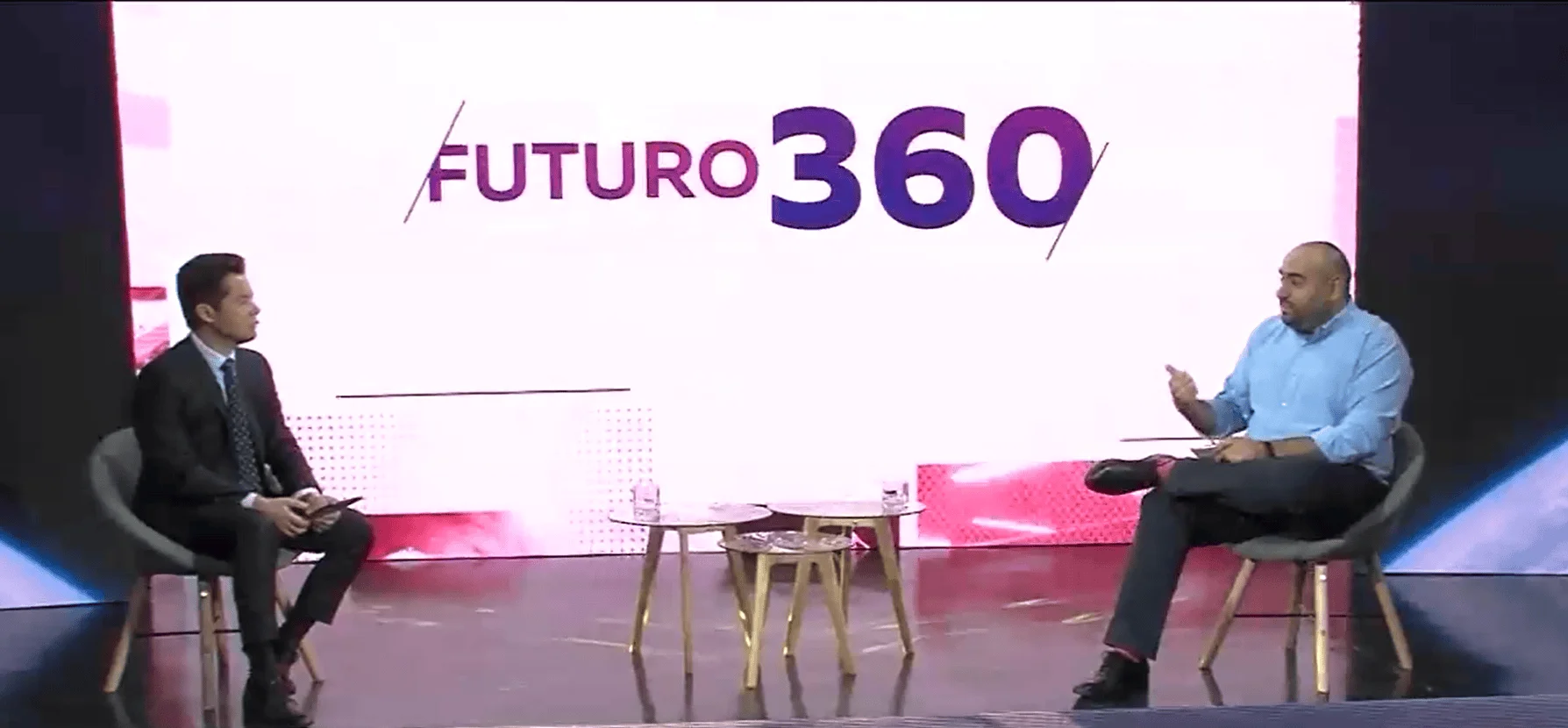 Dos hombres sentados en sillas conversan en el escenario del evento Futuro 360, frente a una pantalla con el logo del programa.