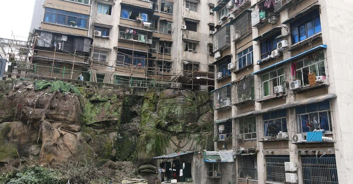 Bloques de apartamentos deteriorados en ciudad china con una gran roca cubierta de musgo y relieves esculpidos en la base.