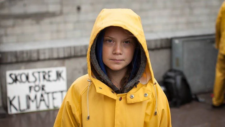 Joven con impermeable amarillo frente a un cartel que dice 'Skolstrejk för Klimatet', huelga escolar por el clima.