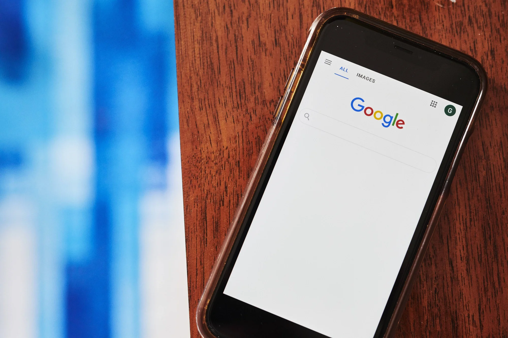 Teléfono inteligente apoyado en una mesa de madera mostrando la página de inicio del buscador Google.