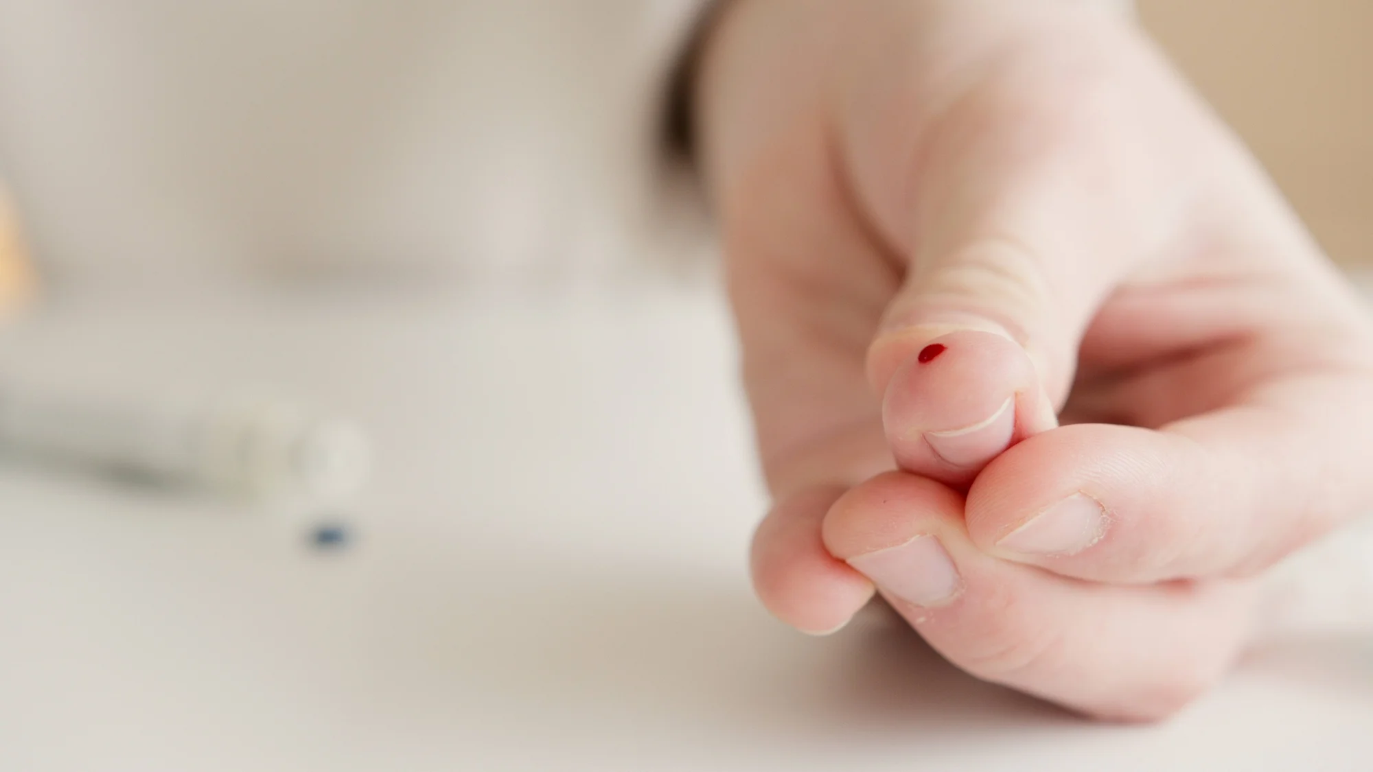 Primer plano de un dedo con una pequeña gota de sangre en la yema, indicando una punción para análisis de glucosa en sangre.