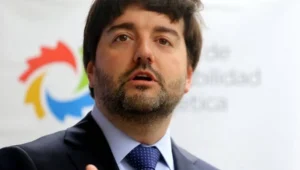 Hombre con traje oscuro y corbata azul habla frente a un logo institucional de colores.
