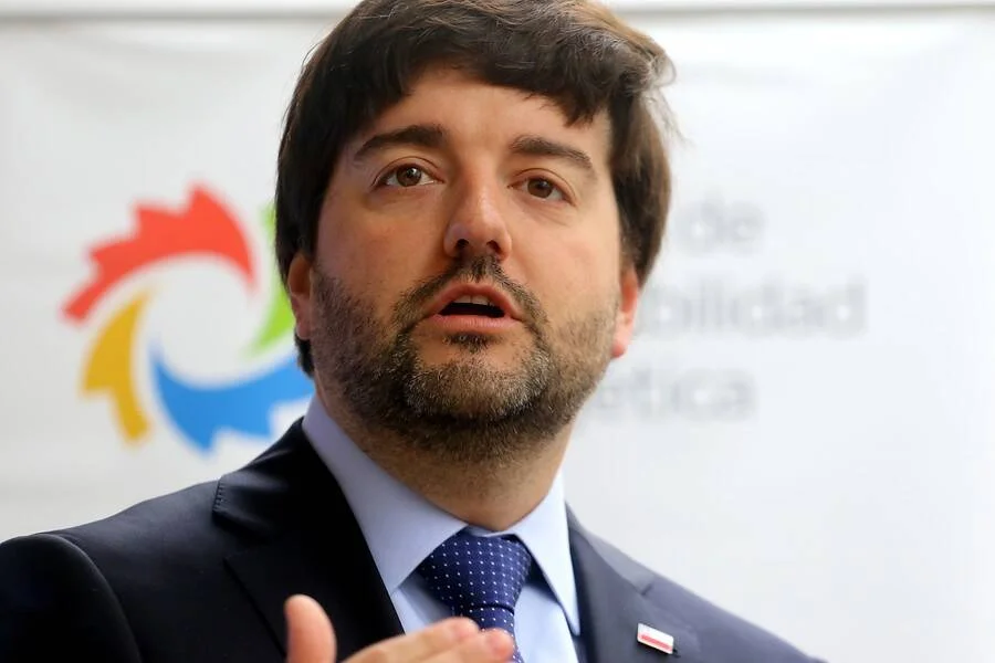 Hombre con traje oscuro y corbata azul habla frente a un logo institucional de colores.