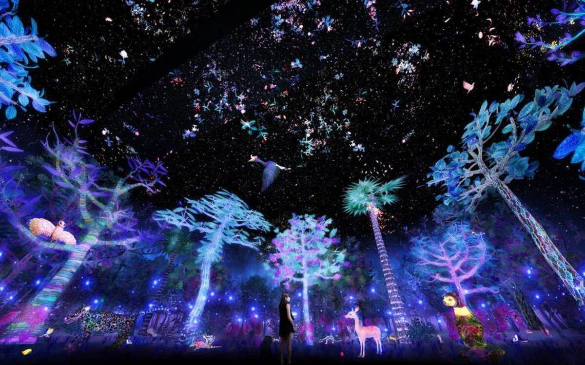 Persona de pie en una instalación inmersiva de arte digital con árboles y animales iluminados en tonos azules y violetas bajo un cielo estrellado.