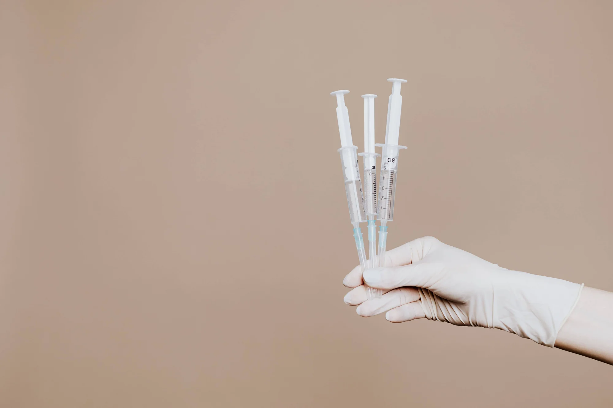 Mano enguantada con látex sostiene tres jeringas médicas vacías sobre fondo beige.