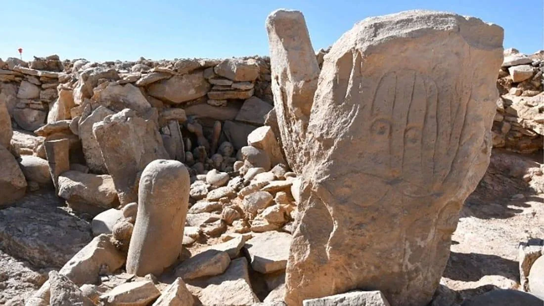 Bloque de piedra con figura humana grabada en un sitio arqueológico desértico rodeado de rocas y muros de piedra.