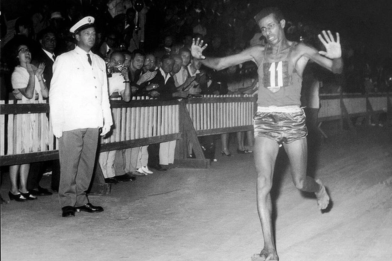 Atleta descalzo con dorsal número 11 cruza la línea de meta con los brazos abiertos ante una multitud que aplaude, en foto histórica en blanco y negro.