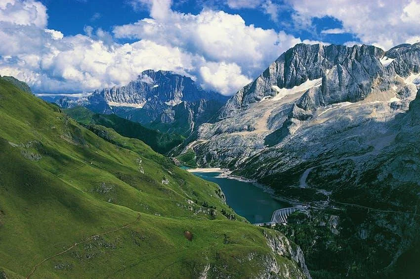 Valle alpino con lago turquesa, presa y picos rocosos nevados bajo cielo nublado en verano.