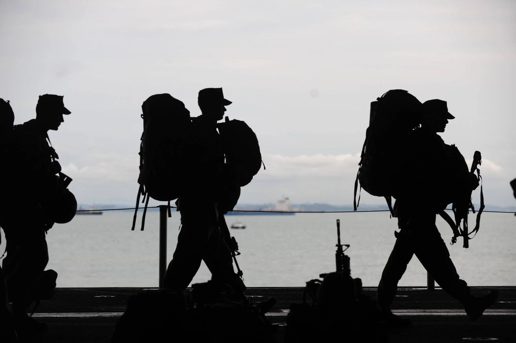 Siluetas de soldados con mochilas militares y armas marchando en fila frente a un horizonte marino.