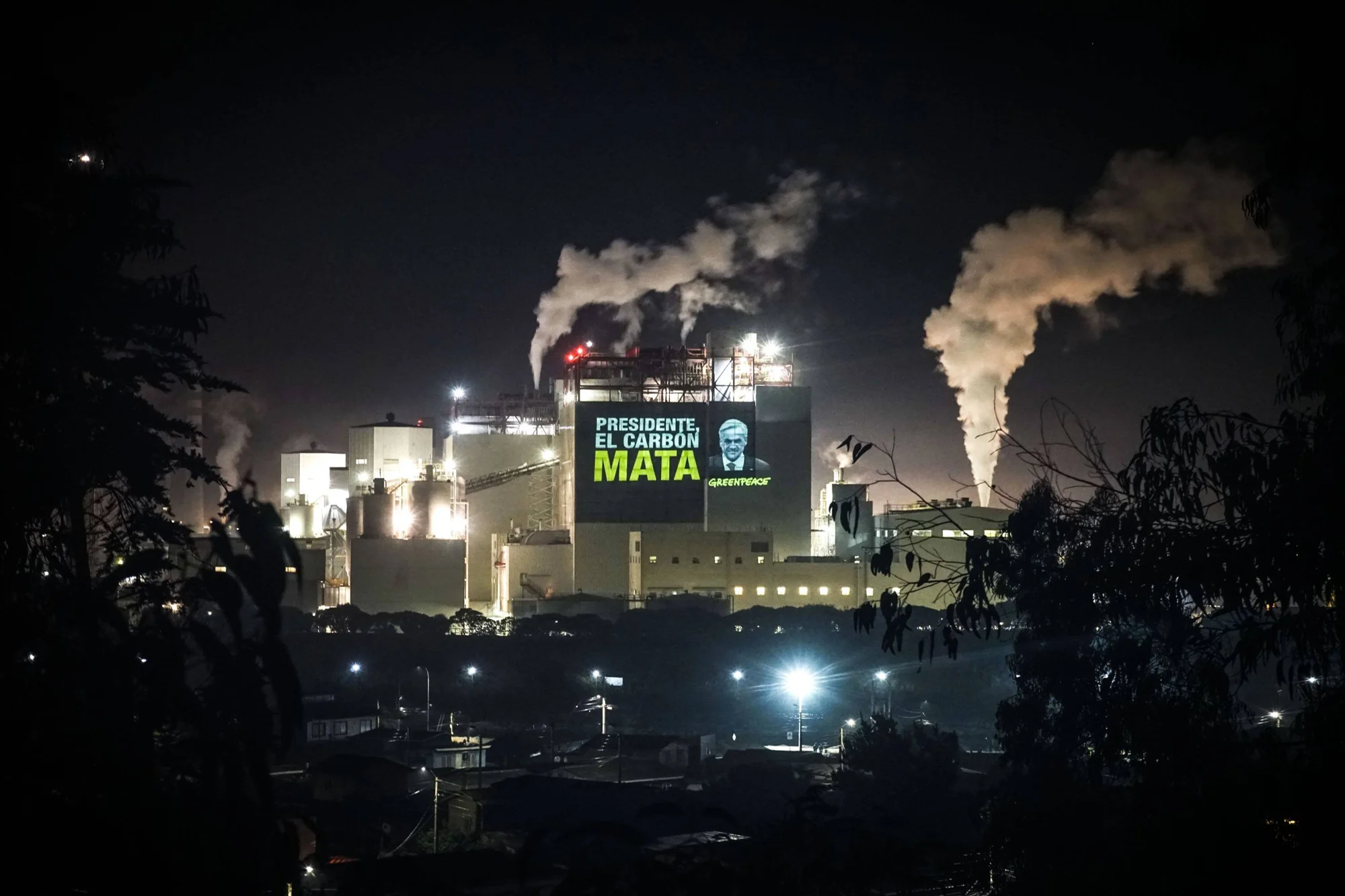 Greenpeace proyecta el mensaje 'Presidente, el carbón mata' sobre una planta industrial que emite humo, durante la noche.