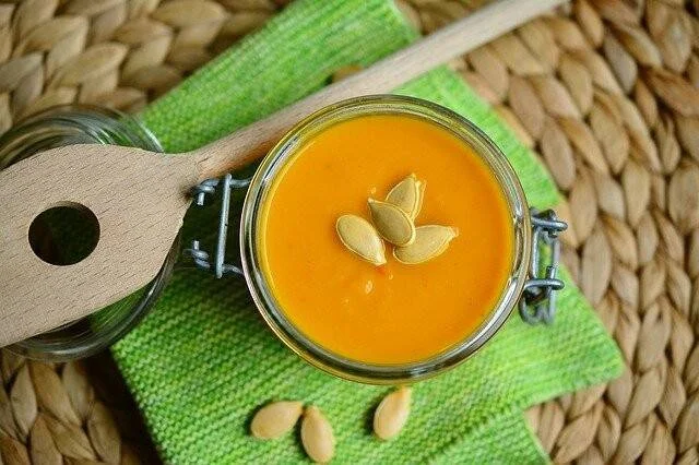 Crema de calabaza naranja en frasco hermético, decorada con semillas de zapallo, junto a una cuchara de madera sobre servilleta verde.