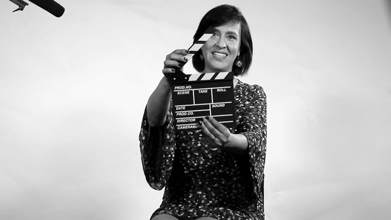 Mujer sonriente sostiene una claqueta de producción cinematográfica frente a cámara, en foto en blanco y negro.